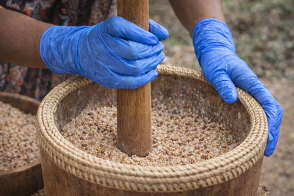 Person using a wooden pestle in a woven container filled with grains, wearing blue gloves.