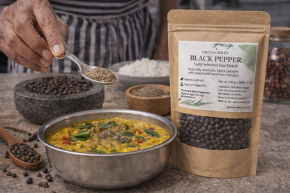 Black pepper being added to a dish with a package of Green Origin Black Pepper on a wooden surface.