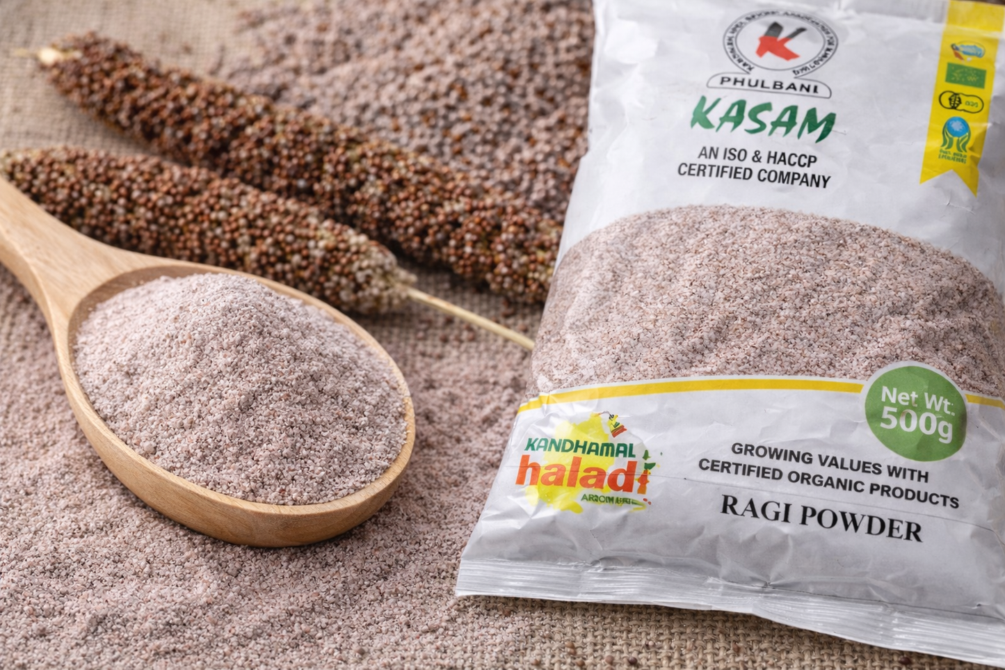 Ragi powder in a wooden spoon with a package labeled 'Kasam' and 'Haladi' on a textured surface.